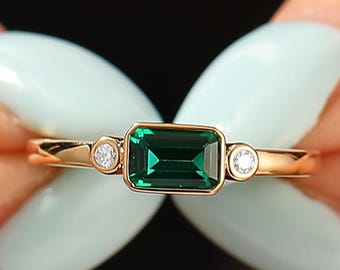 May include: A gold ring featuring a rectangular emerald-cut green gemstone, flanked by two smaller, round, clear gemstones. The ring is held up by a person with light blue nail polish.