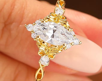Op de afbeelding: Een gouden ring met een heldere, marquise-geslepen edelsteen in het midden, omringd door kleinere, ronde, heldere edelstenen. De ring heeft een sierlijk ontwerp en een gedraaide band. De ring wordt tussen de vingers gehouden.