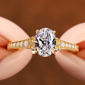 Puede incluir: Un anillo de oro con una piedra central de diamante de talla ovalada. La banda presenta peque&ntilde;as piedras de acento redondas y filigranas decorativas. El anillo se sostiene sobre un fondo marr&oacute;n borroso.