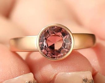 May include: A rose gold ring with a round, faceted, pink gemstone. The ring has a simple band and a bezel setting. The gemstone is the focal point, catching the light and reflecting a warm, rosy hue. The ring is a piece of jewelry.