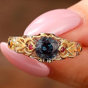 Könnte beinhalten: Ein Goldring mit einem zentralen, runden, dunkelblauen Edelstein, der von zwei kleinen roten Steinen flankiert wird. Das Band ist mit einem komplizierten, erhabenen Filigran-Metallmuster gestaltet, das sich in einem durchgehenden Design um die Fassung legt.