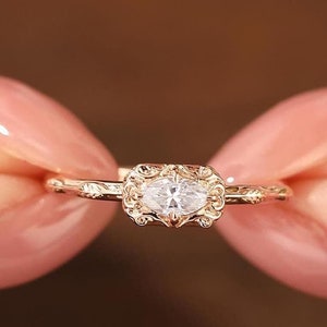 Pode incluir: Um delicado anel de ouro com um pequeno diamante oval engastado em um design ornamentado e detalhado.