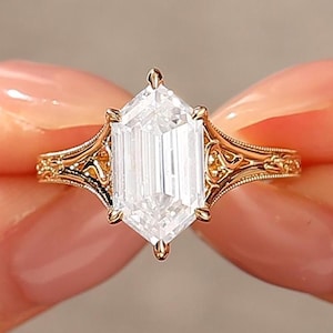 Op de afbeelding: Een gouden ring met een heldere, smaragdgeslepen edelsteen. De ring heeft een sierlijk filigraanpatroon op de band en rond de edelsteen setting. De ring wordt tussen twee vingers gehouden tegen een neutrale achtergrond.