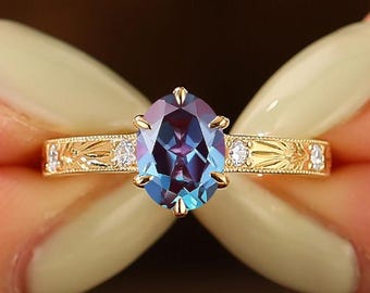 May include: A gold ring featuring an oval-cut color-changing gemstone in the center, held by six prongs. The band is engraved with a leaf-like pattern and accented with small, round, clear stones on either side of the central setting.