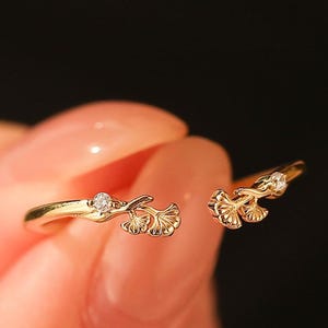 May include: A gold-colored ring with a split design, featuring delicate ginkgo leaf details and small, sparkling gemstones. The ring is held up against a blurred background, showcasing its intricate design and elegant style.