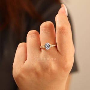 Puede incluir: Un anillo de compromiso de oro con una piedra central de diamante de talla ovalada. La banda presenta peque&ntilde;as piedras de acento y detalles delicados. El anillo est&aacute; en el dedo de una persona.