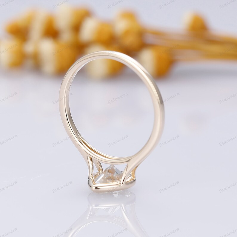 Pu&ograve; includere: Un anello di fidanzamento in oro con un diamante di forma quadrata incastonato a griffe. L'anello &egrave; su una superficie bianca.