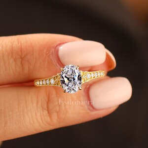 Puede incluir: Un anillo de compromiso de oro con una gran piedra central de diamante de talla ovalada. La banda tambi&eacute;n es de oro y presenta diamantes de acento m&aacute;s peque&ntilde;os. El anillo se sostiene contra un fondo borroso.