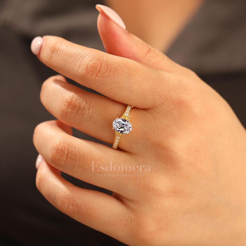 Puede incluir: Un anillo de oro con un gran diamante central de talla ovalada. La banda tambi&eacute;n est&aacute; adornada con diamantes m&aacute;s peque&ntilde;os. El anillo est&aacute; en el dedo de una persona. La palabra "Esdomera" est&aacute; impresa en la imagen.