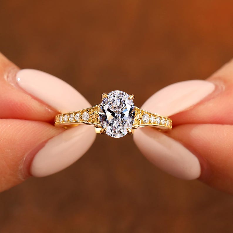Puede incluir: Un anillo de oro con una piedra central de diamante de talla ovalada. La banda presenta peque&ntilde;as piedras de acento redondas y filigranas decorativas. El anillo se sostiene sobre un fondo marr&oacute;n borroso.