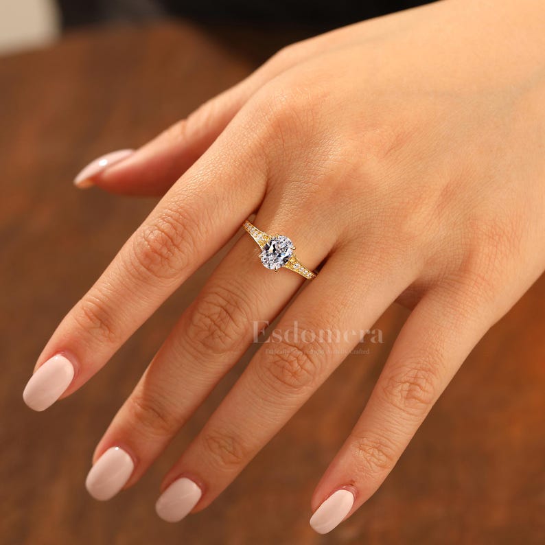 Puede incluir: Un anillo de oro con un gran diamante central de talla ovalada. La banda tambi&eacute;n est&aacute; adornada con diamantes m&aacute;s peque&ntilde;os. El anillo est&aacute; en una mano con u&ntilde;as cuidadas. La palabra "Esdomera" es visible.