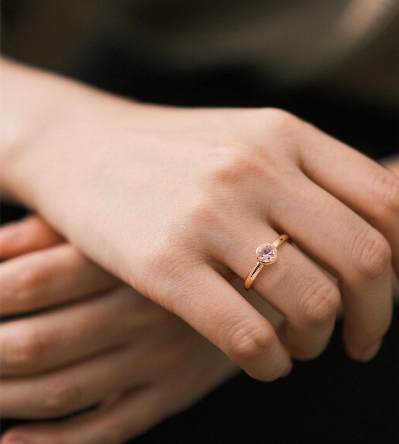 A delicate solid yellow gold ring with a bezel setting featuring a round, light pink gemstone.