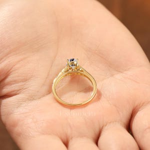 Puede incluir: Un anillo de oro con una piedra preciosa redonda y transparente engastada en un engaste de cuatro puntas. La banda del anillo tambi&eacute;n es dorada y tiene peque&ntilde;as piedras transparentes. El anillo se sostiene en una mano.