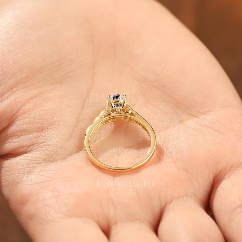 Puede incluir: Un anillo de oro con una piedra preciosa redonda y transparente engastada en un engaste de cuatro puntas. La banda del anillo tambi&eacute;n es dorada y tiene peque&ntilde;as piedras transparentes. El anillo se sostiene en una mano.