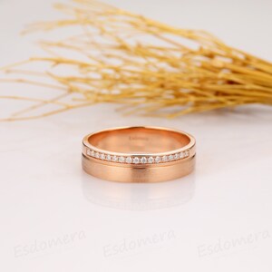 May include: A rose gold wedding band with a row of small diamonds set in a channel setting. The band has a brushed finish.