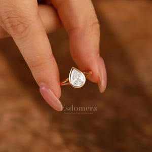 Pu&ograve; includere: Un anello di fidanzamento in oro con un diamante a forma di pera. L'anello &egrave; tenuto da una mano con unghie curate.