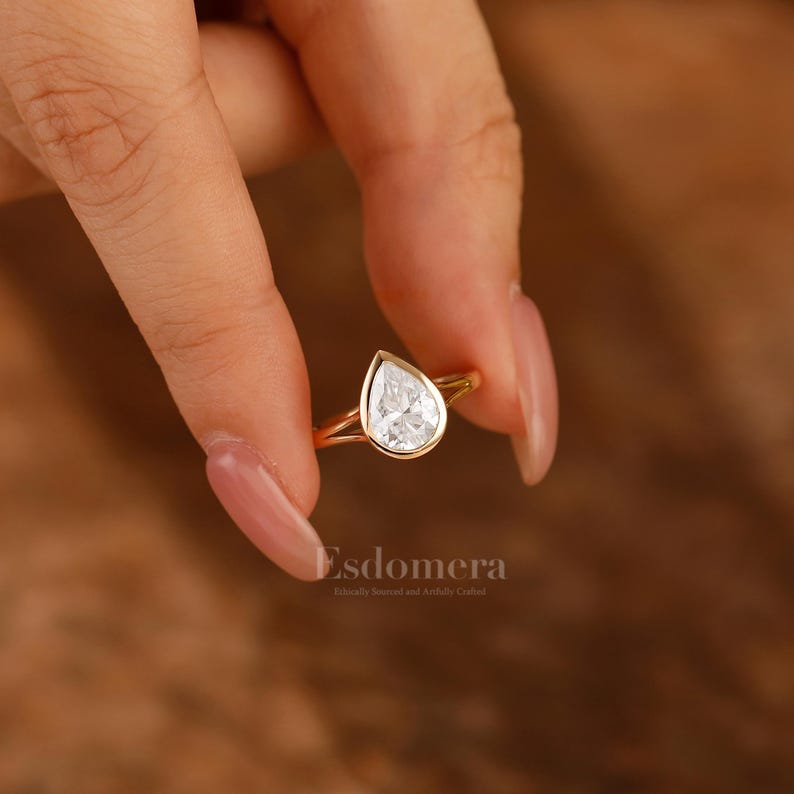 Pu&ograve; includere: Un anello di fidanzamento in oro con un diamante a forma di pera. L'anello &egrave; tenuto da una mano con unghie curate.