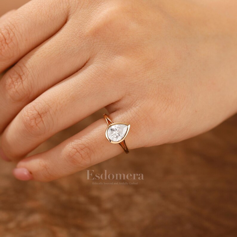 Pu&ograve; includere: Un anello d'oro con una pietra preziosa trasparente a forma di pera. L'anello &egrave; su un dito.