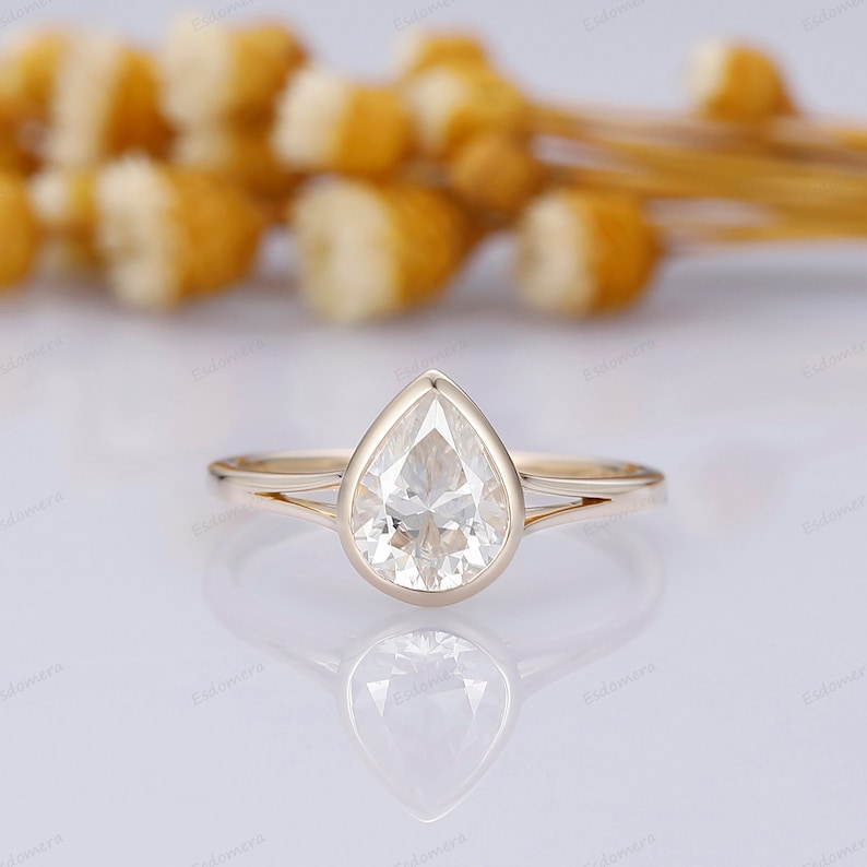 Pu&ograve; includere: Un anello di fidanzamento con un diamante a forma di pera incastonato in una fascia d'oro. L'anello ha un design semplice con un solo diamante al centro.