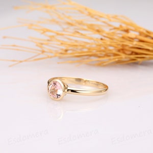 May include: A delicate gold ring with a round, pale pink gemstone set in a bezel setting. The ring is simple and elegant.