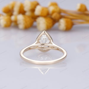 Pu&ograve; includere: Un anello di fidanzamento in oro con un diamante a forma di pera. L'anello &egrave; su una superficie bianca con uno sfondo sfocato di fiori gialli.