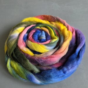 Puede incluir: Una madeja de lana multicolor vibrante, dispuesta en espiral. La lana muestra un espectro de colores, incluyendo amarillo, rosa, azul, verde y morado. La mecha está bien enrollada, creando un diseño texturizado visualmente atractivo, perfecto para proyectos de arte textil.