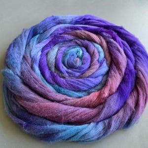 May include: A circular arrangement of dyed fibre in shades of blue, purple, and pink. The fibre is wound in a spiral, creating a visually interesting pattern. The texture appears soft and fluffy, ideal for textile crafts.
