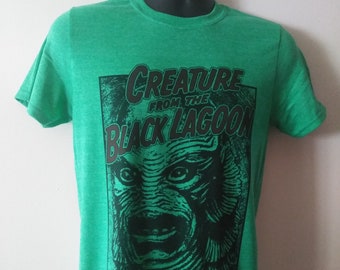 May include: Green t-shirt with a black and white graphic print of the Creature from the Black Lagoon. The graphic features the creature's face with a menacing expression.
