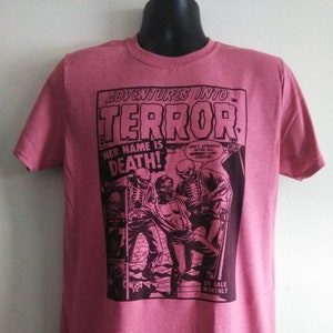 May include: A pink t-shirt featuring a comic book-style graphic with the words "ADVENTURES INTO TERROR." The graphic includes the text "HER NAME IS DEATH!" and illustrations of skeletons and a person. The shirt is displayed on a mannequin.