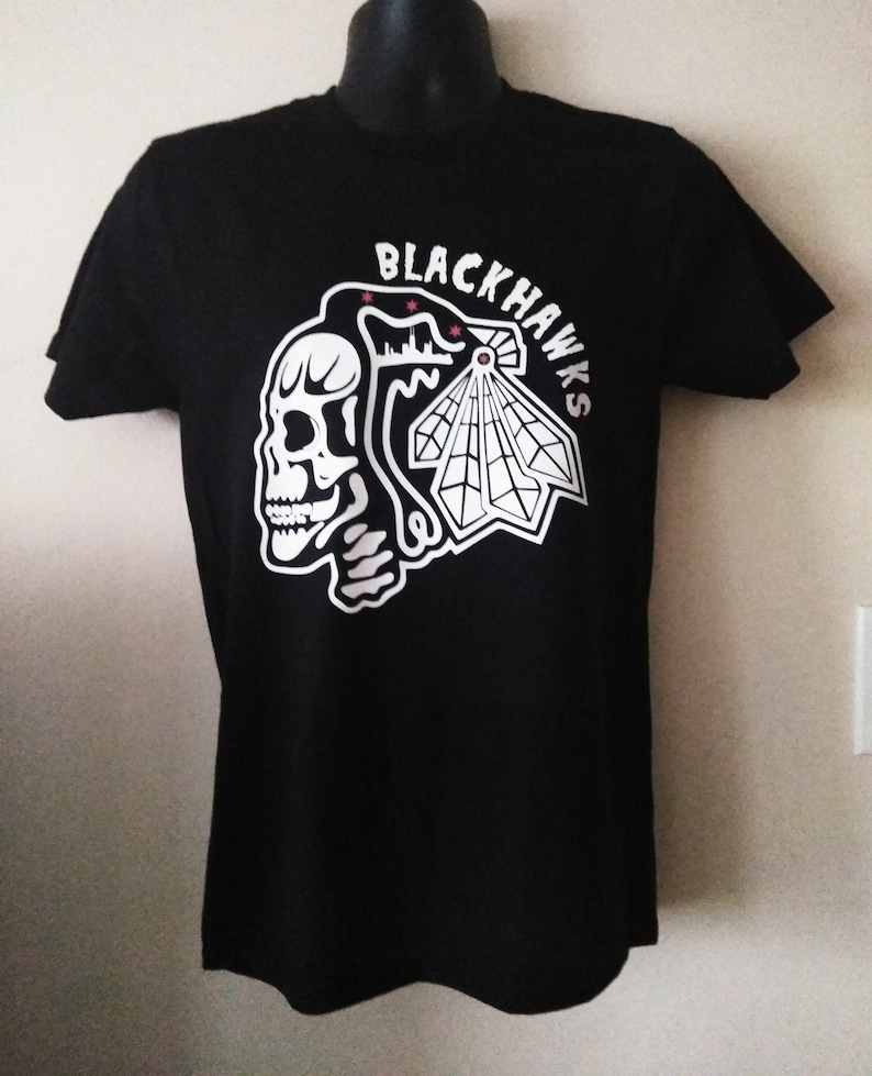 May include: Black t-shirt with a white graphic of a skull and a stylized Chicago Blackhawks logo. The logo includes the text "Blackhawks" and three red stars.