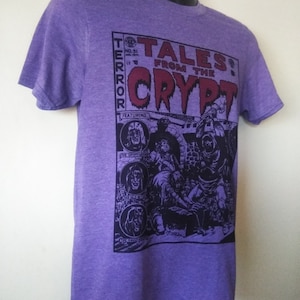 May include: Purple t-shirt with a graphic print of a comic book cover featuring the text "Tales from the Crypt" and a black and white illustration of a group of monsters. The comic book cover also features the text "Terror" and "Featuring".