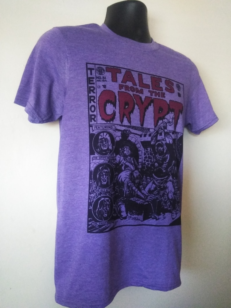 May include: Purple t-shirt with a graphic print of a comic book cover featuring the text "Tales from the Crypt" and a black and white illustration of a group of monsters. The comic book cover also features the text "Terror" and "Featuring".