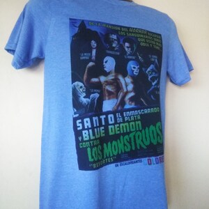 May include: Light blue t-shirt featuring a vintage movie poster design. The poster includes text in Spanish, images of masked wrestlers, and classic horror movie characters. The shirt has short sleeves and a crew neck.