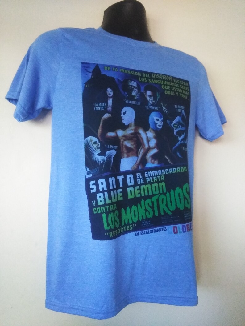 May include: Light blue t-shirt featuring a vintage movie poster design. The poster includes text in Spanish, images of masked wrestlers, and classic horror movie characters. The shirt has short sleeves and a crew neck.