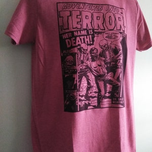May include: A maroon t-shirt featuring a comic book-style graphic. The graphic reads "ADVENTURES INTO TERROR" with the text "HER NAME IS DEATH!" and illustrations of skeletons and a person. The shirt also has the text "ON SALE MONTHLY".