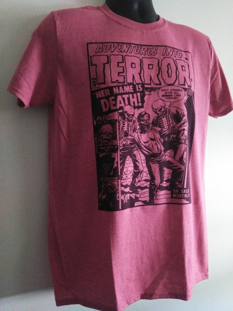 May include: A maroon t-shirt featuring a comic book-style graphic. The graphic reads "ADVENTURES INTO TERROR" with the text "HER NAME IS DEATH!" and illustrations of skeletons and a person. The shirt also has the text "ON SALE MONTHLY".