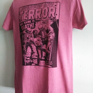 May include: Pink t-shirt featuring a comic book cover design. The graphic reads "ADVENTURES INTO TERROR!" with the text "HER NAME IS DEATH!" and a vintage illustration of a horror scene. The shirt has short sleeves and a crew neck.