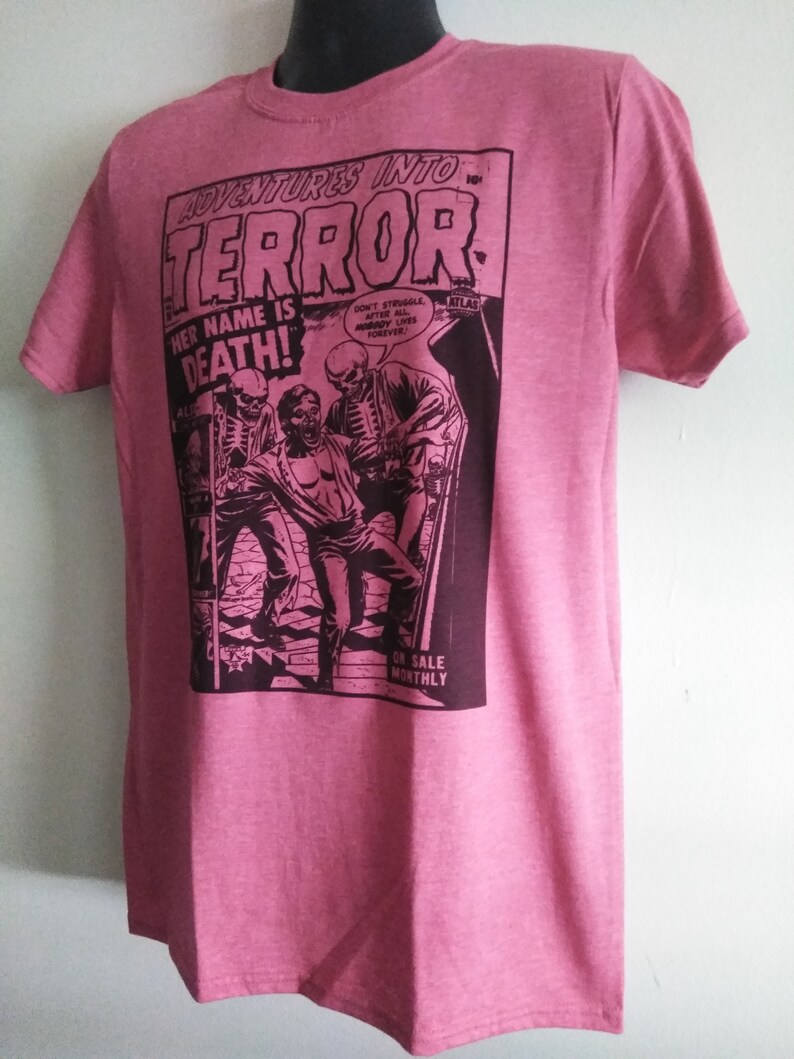 May include: Pink t-shirt featuring a comic book cover design. The graphic reads "ADVENTURES INTO TERROR!" with the text "HER NAME IS DEATH!" and a vintage illustration of a horror scene. The shirt has short sleeves and a crew neck.