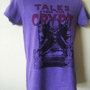 May include: Purple t-shirt with a graphic print of a character from the Tales from the Crypt television series. The character is sitting on a throne with a book in his lap. The text "Tales from the Crypt" is printed in red above the character.