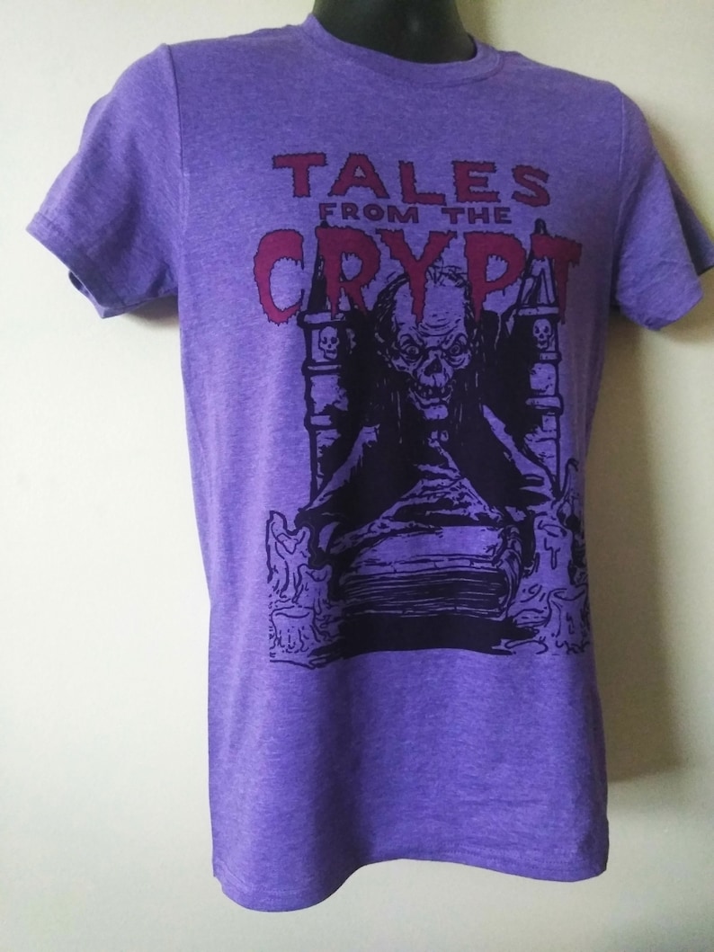 May include: Purple t-shirt with a graphic print of a character from the Tales from the Crypt television series. The character is sitting on a throne with a book in his lap. The text "Tales from the Crypt" is printed in red above the character.