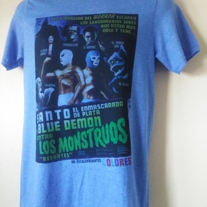 May include: Light blue t-shirt featuring a vintage movie poster design. The poster includes images of monsters and wrestlers, with text in Spanish. The shirt has short sleeves and a crew neck.