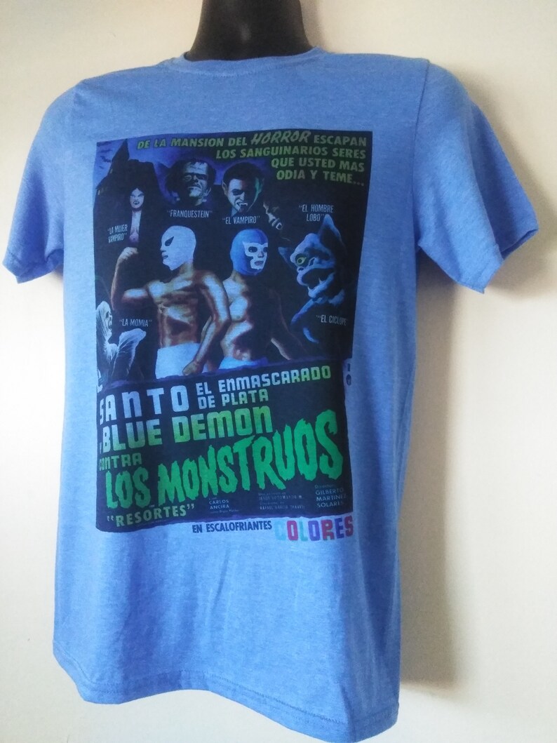 May include: Light blue t-shirt featuring a vintage movie poster design. The poster includes images of monsters and wrestlers, with text in Spanish. The shirt has short sleeves and a crew neck.