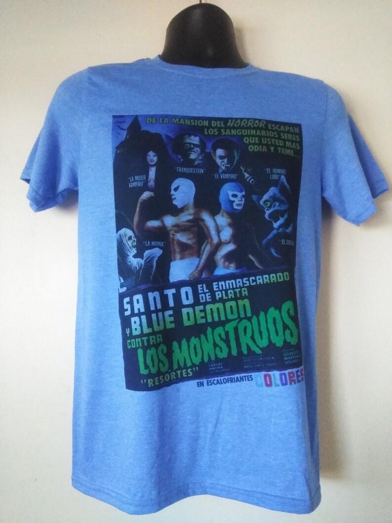 May include: Light blue t-shirt featuring a movie poster design. The poster includes images of masked wrestlers and classic horror movie characters. Text on the shirt includes "SANTO EL ENMASCARADO DE PLATA BLUE DEMON CONTRA LOS MONSTRUOS".