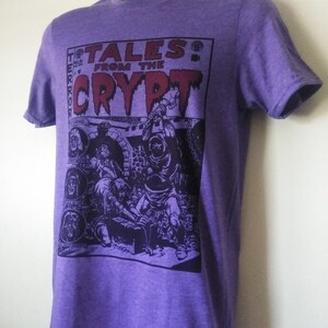May include: Purple t-shirt featuring a graphic print of a comic book cover with the text "Tales from the Crypt" and a black and white illustration of a scene from the comic book.