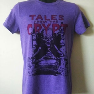 May include: Purple t-shirt with a graphic print of a spooky figure sitting on a book. The text "Tales from the Crypt" is printed in red above the figure.
