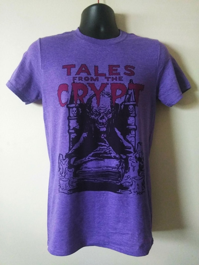 May include: Purple t-shirt with a graphic print of a spooky figure sitting on a book. The text "Tales from the Crypt" is printed in red above the figure.