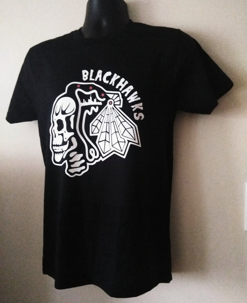 May include: Black t-shirt with a white graphic of a skull and a stylized Native American headdress with the text "BLACKHAWKS" around it.