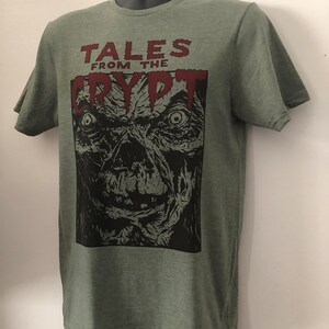 May include: A green t-shirt with a black and white illustration of a monster's face. The text "Tales from the Crypt" is printed in red above the illustration.