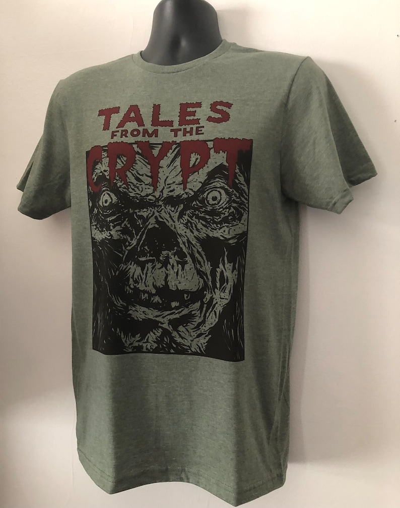 May include: A green t-shirt with a black and white illustration of a monster's face. The text "Tales from the Crypt" is printed in red above the illustration.