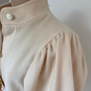 May include: A light peach-colored coat with a high collar and a single large button. The coat features a classic design with gathered sleeves, offering a vintage aesthetic. The fabric appears soft and textured.